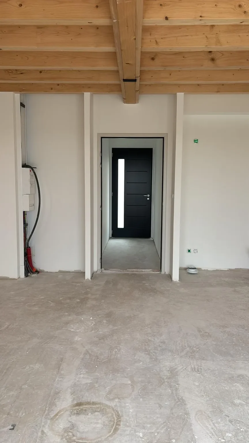 Intérieur d'une maison en construction avec des poutres en bois apparentes, un sol en béton brut et une porte d'entrée noire moderne.