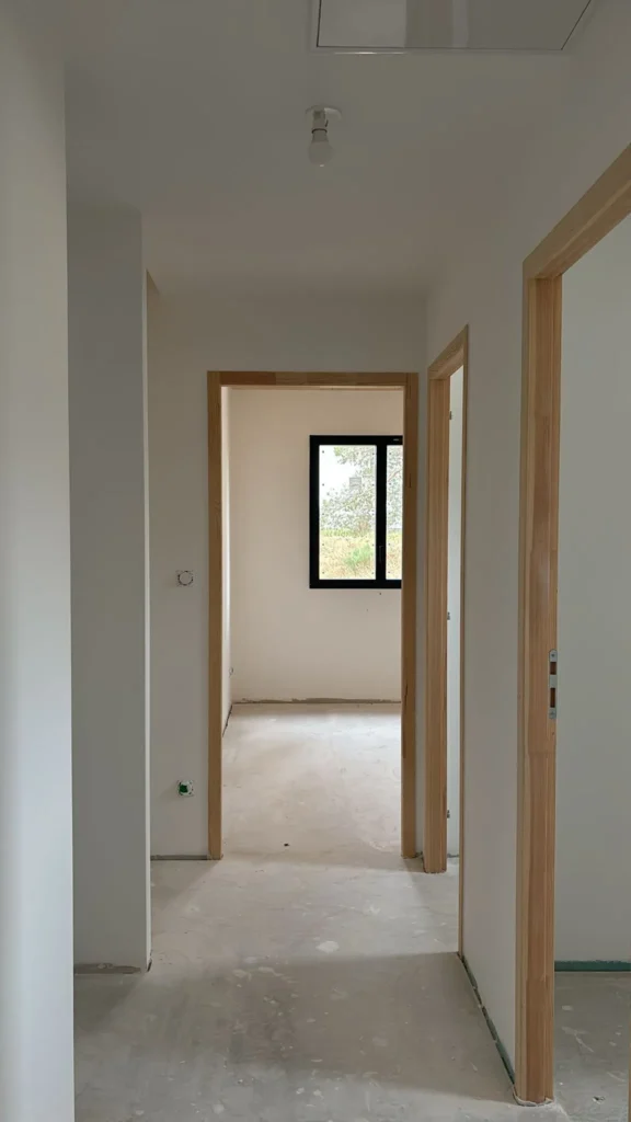 Couloir d'un bâtiment en construction avec plusieurs portes en bois ouvrant sur des pièces vides, dont une avec une fenêtre