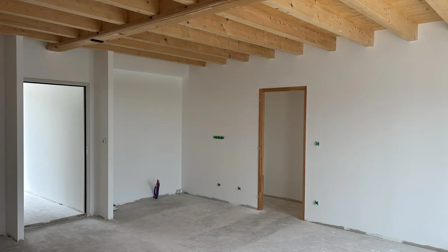 Intérieur d'une maison en construction, avec un plafond en poutres de bois apparentes, murs blancs, sol en béton et deux portes.