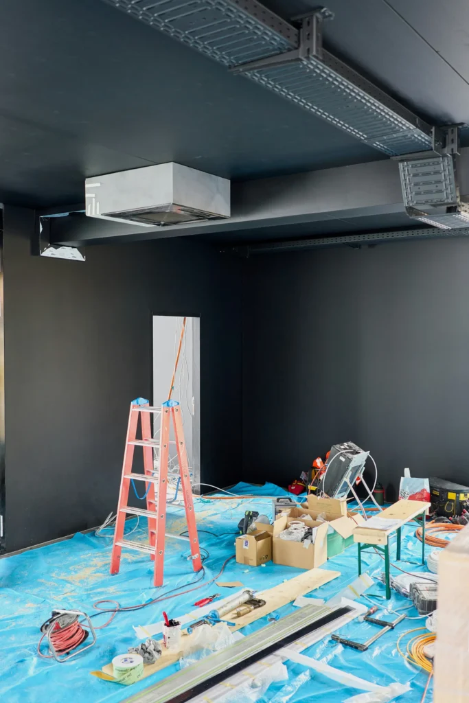Chantier intérieur avec murs sombres, plafond avec chemins de câbles, un escalier rouge et du matériel de construction au sol.