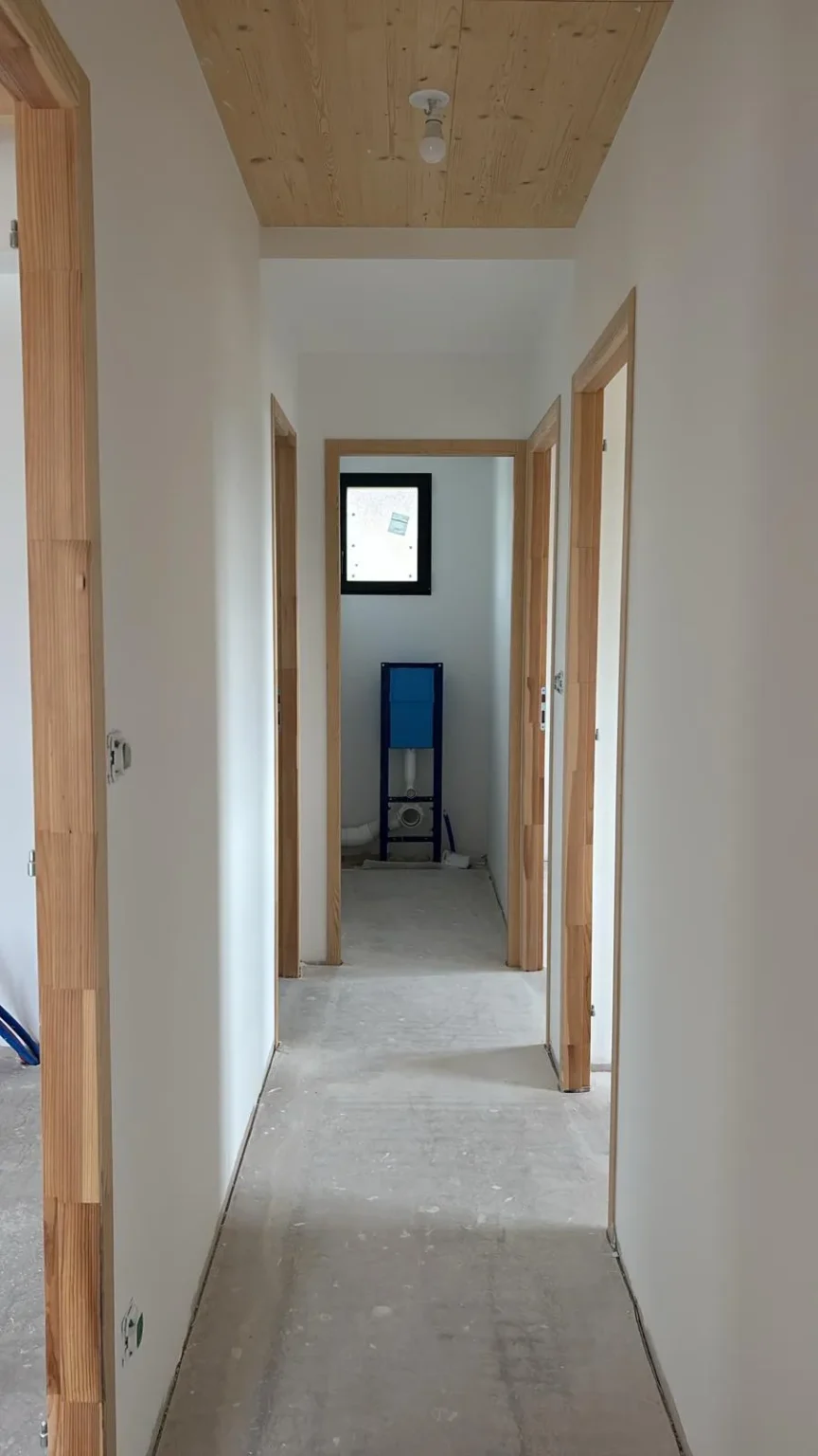 Couloir en construction avec murs blancs, boiseries en bois clair, sol en béton brut et un élément de chasse d'eau suspendue au fond.