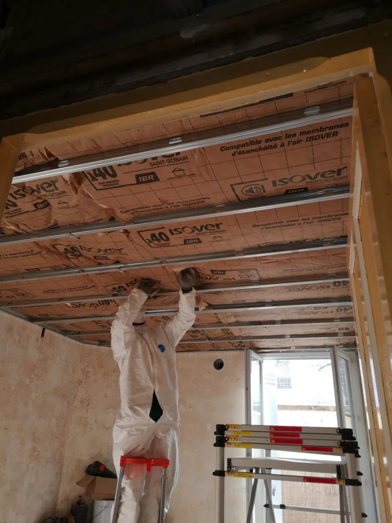 Un ouvrier en combinaison blanche installe un isolant sur le plafond d'une pièce en cours de rénovation.