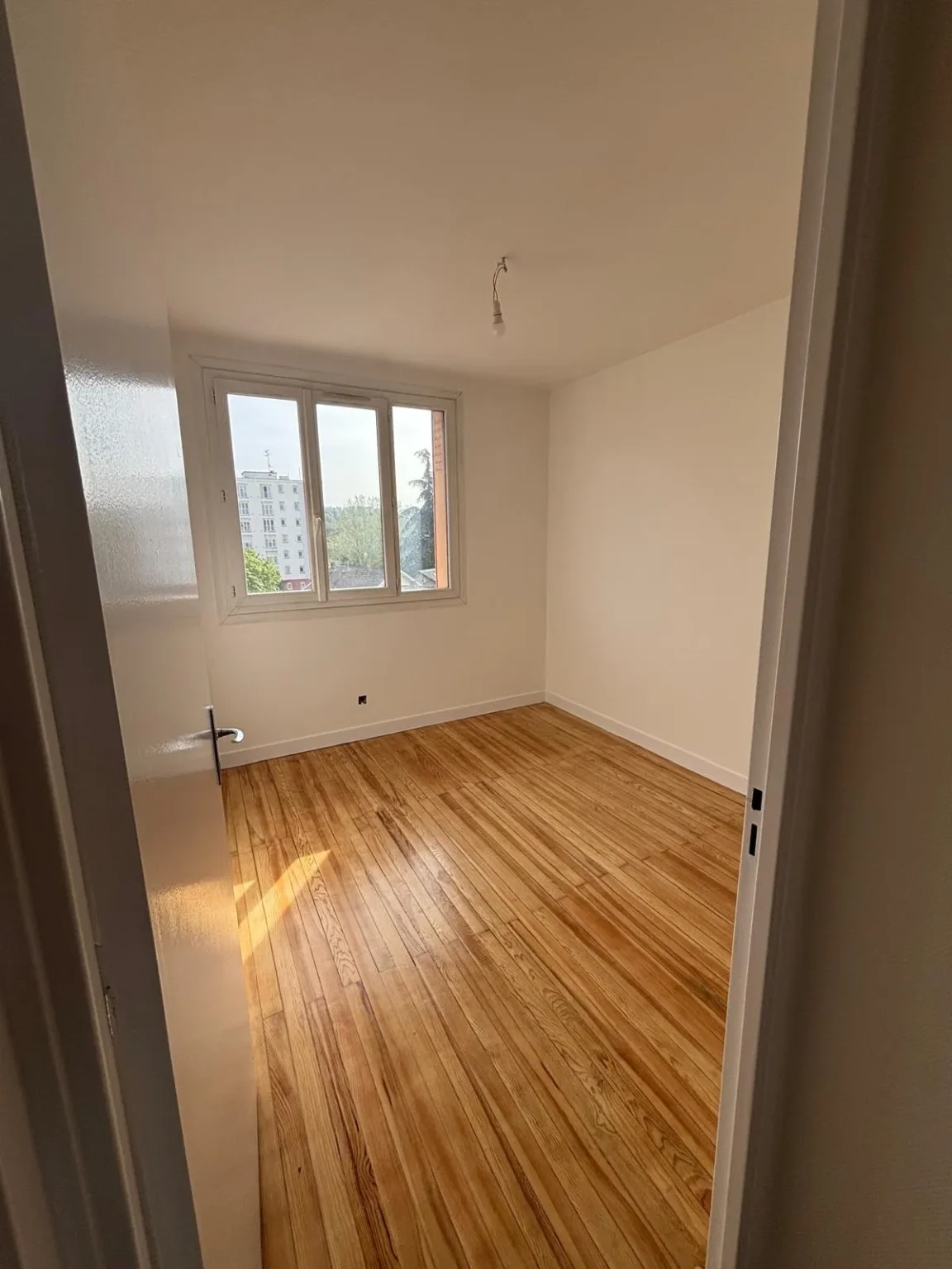 Chambre avec parquet en bois clair, murs blancs et une grande fenêtre donnant sur un immeuble