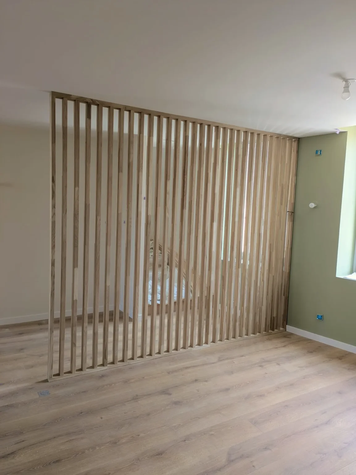 Une cloison ajourée en lattes de bois sépare un salon clair au parquet bois d'une pièce adjacente voilée.