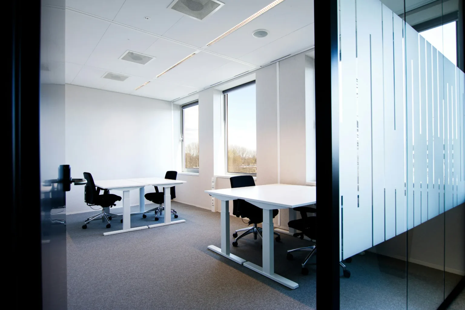Bureau moderne avec deux tables blanches disposées parallèlement, chacune avec deux chaises de bureau noires. Les murs sont blancs, le sol est gris et deux grandes fenêtres éclairent la pièce.