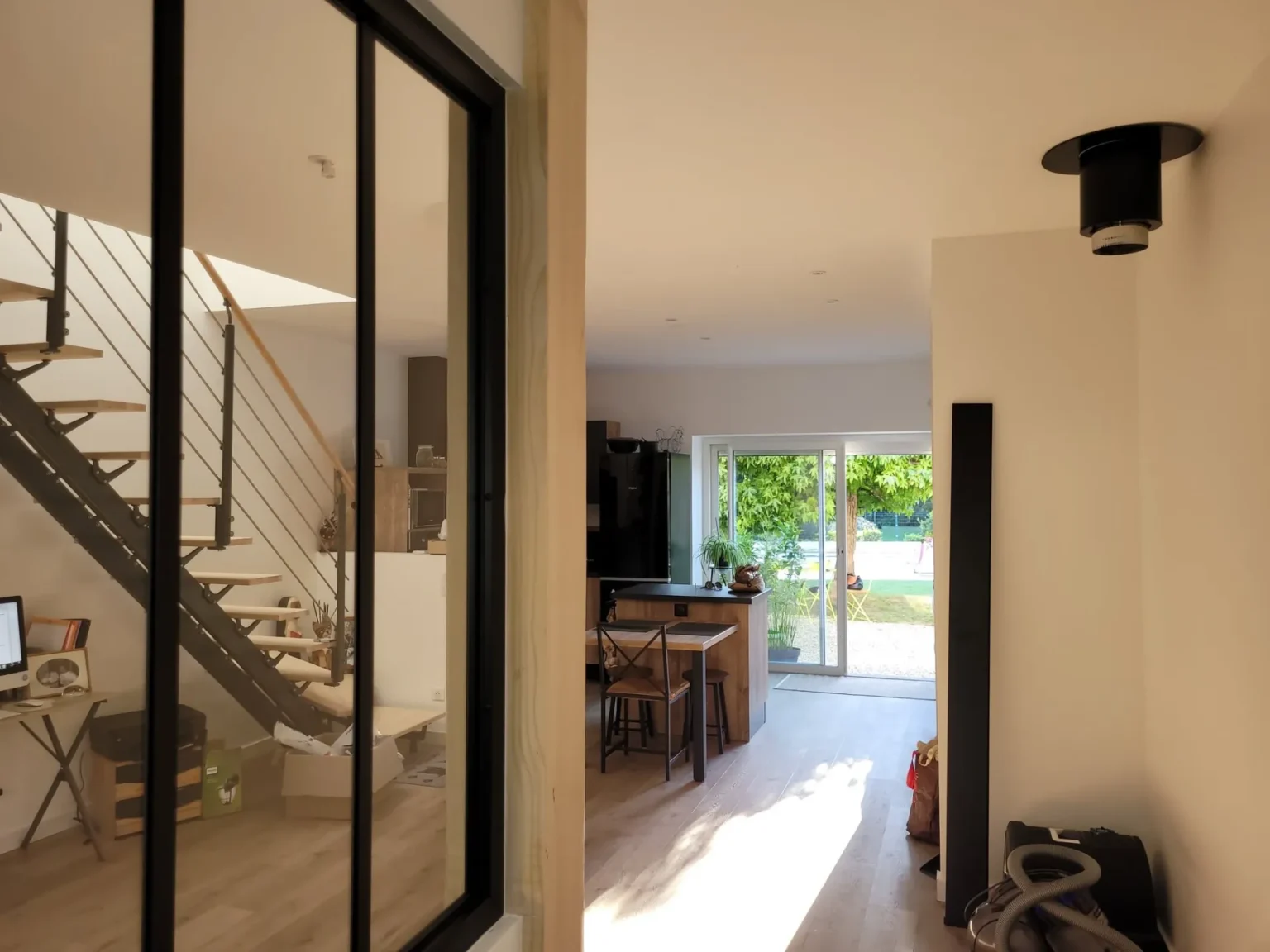 Vue intérieure d'une maison moderne avec escalier, cuisine ouverte et jardin visible par de larges baies vitrées.