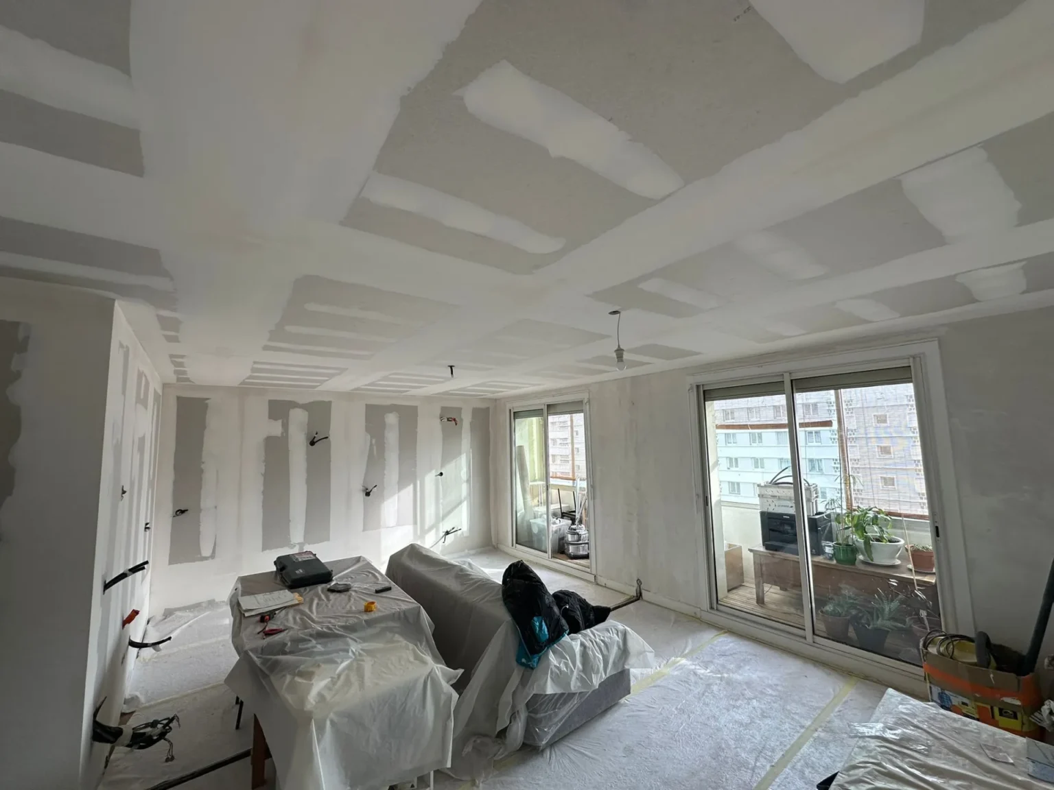 Chantier intérieur : murs et plafond en cours de finition avec plaques de plâtre, mobilier protégé, et grande baie vitrée donnant sur l'extérieur.