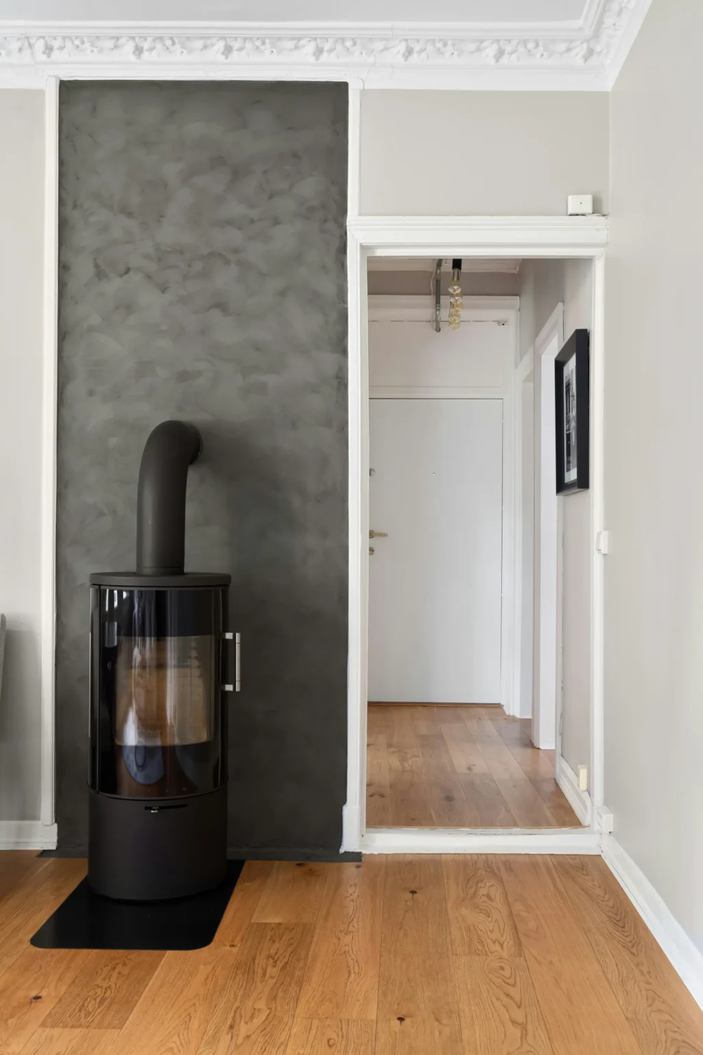 Poêle à bois moderne noir devant un mur texturé gris, avec un couloir blanc menant à une porte blanche en arrière-plan.