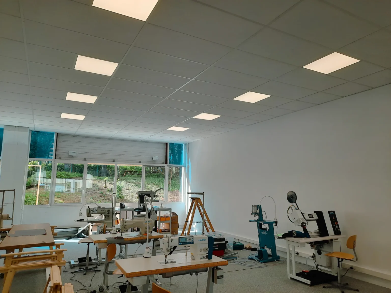 Atelier de couture avec plusieurs machines à coudre, tables de travail et équipements, éclairé par des néons au plafond.