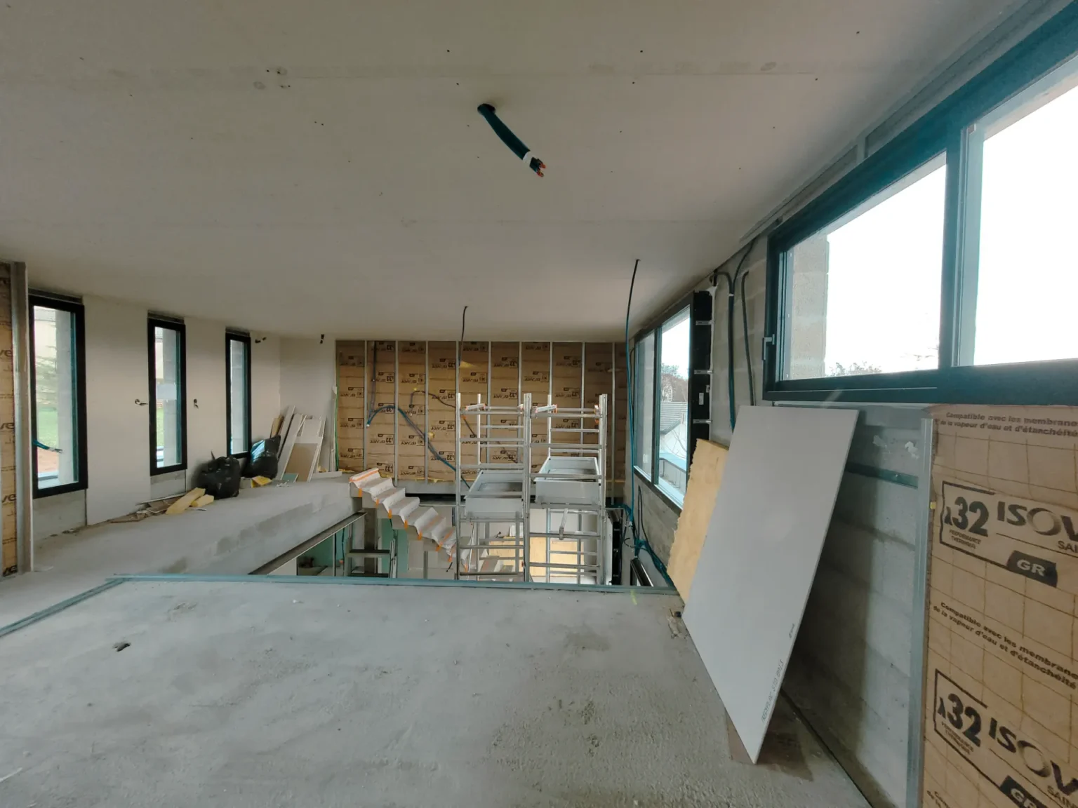 Chantier intérieur d'un grand espace baigné de lumière naturelle, avec une structure d'escalier en cours, des fenêtres et des matériaux de construction.