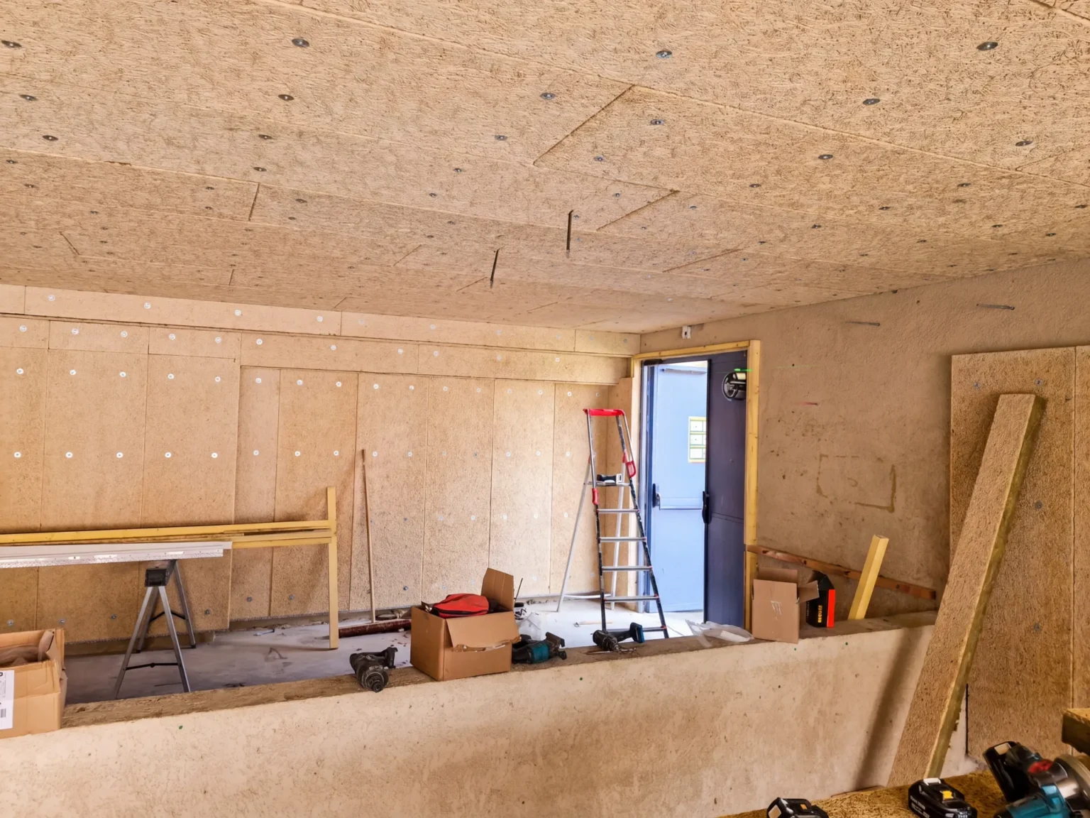 Chantier intérieur : murs et plafond recouverts de panneaux de bois clair, outils et matériaux au sol, porte bleue ouverte