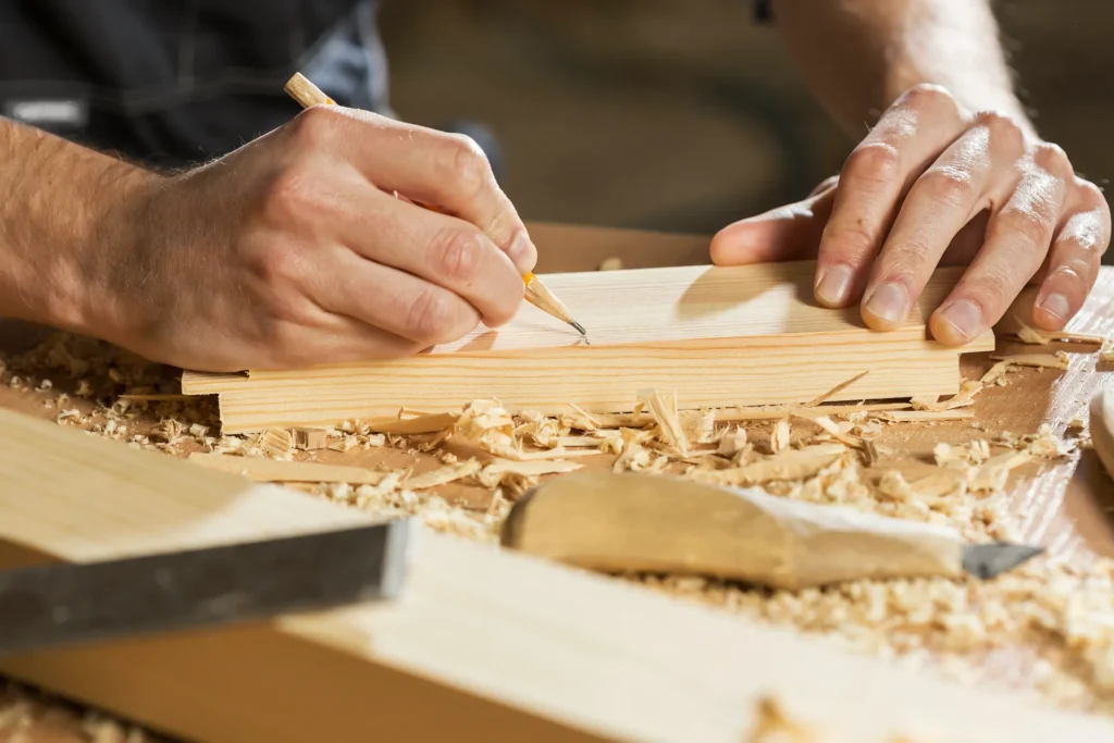 Un menuisier utilise un crayon pour marquer une pièce de bois, entouré de copeaux de bois sur un établi.