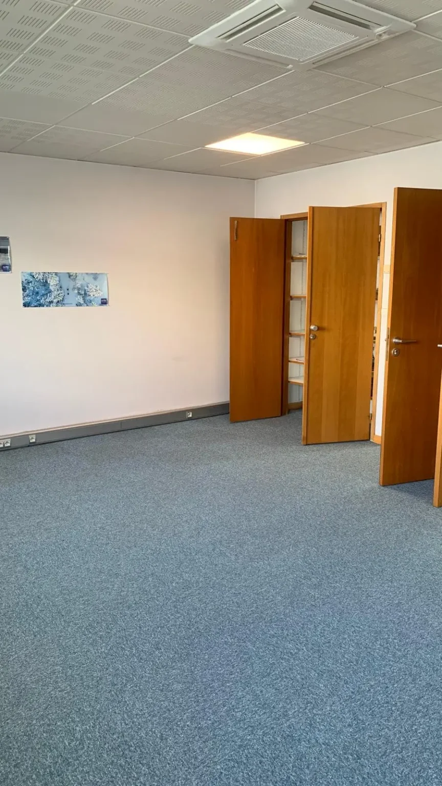 Vue d'un bureau vide avec un tapis bleu, un plafond blanc avec des dalles acoustiques et des portes en bois ouvertes menant à d'autres pièces.
