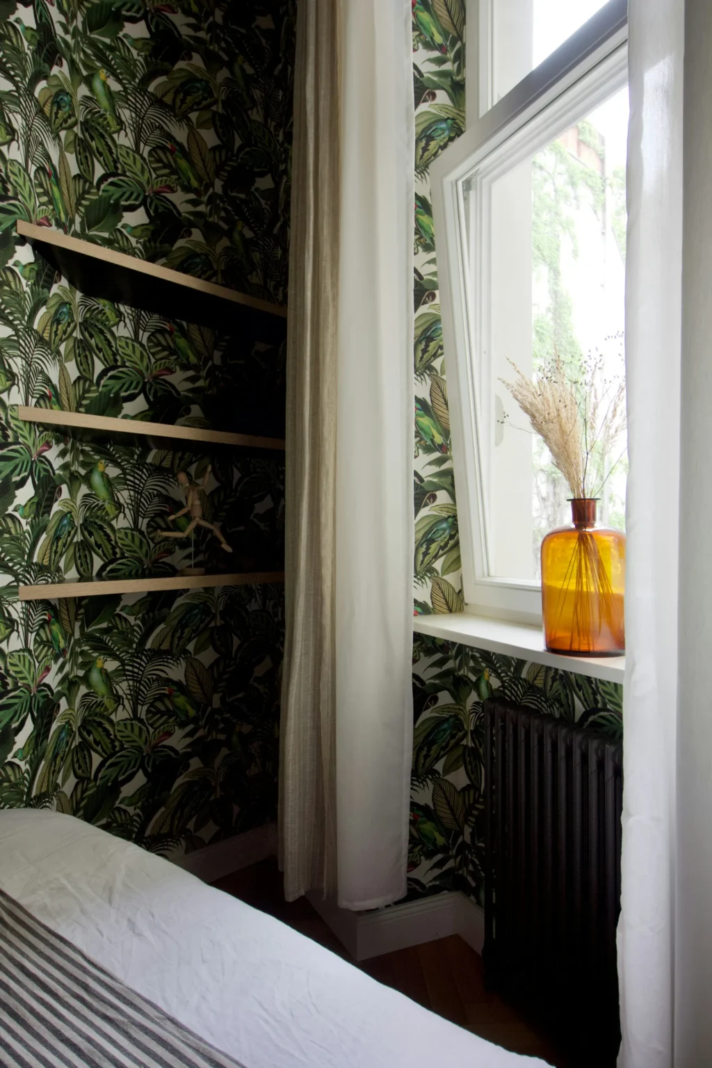 Chambre avec papier peint tropical, étagères en bois, rideaux blanc cassé, et un vase ambré sur le rebord de la fenêtre.