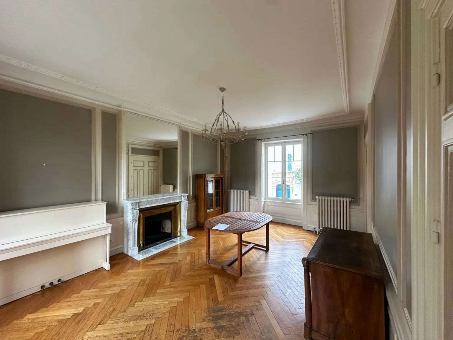 Salon avec parquet en chevrons, cheminée marbre, piano blanc, grande table et buffet en bois.