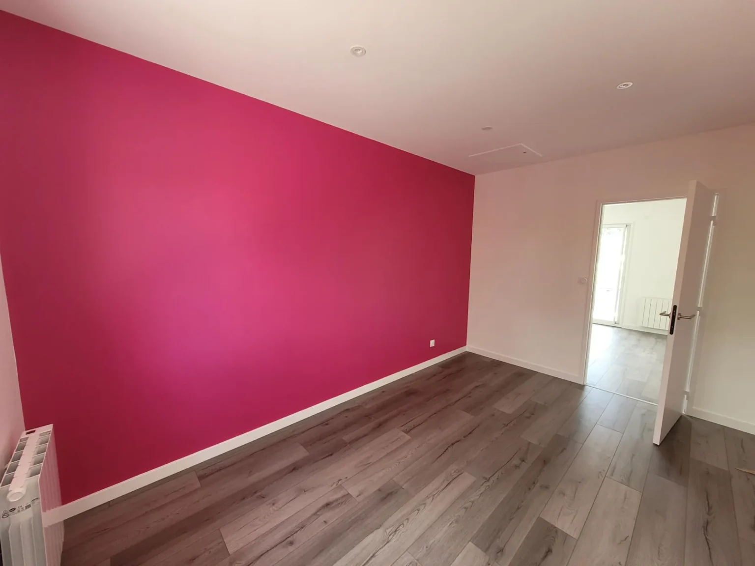 Gros plan sur un mur rose vif peint, dans une pièce avec un sol en parquet gris et une porte blanche ouverte sur une autre pièce.