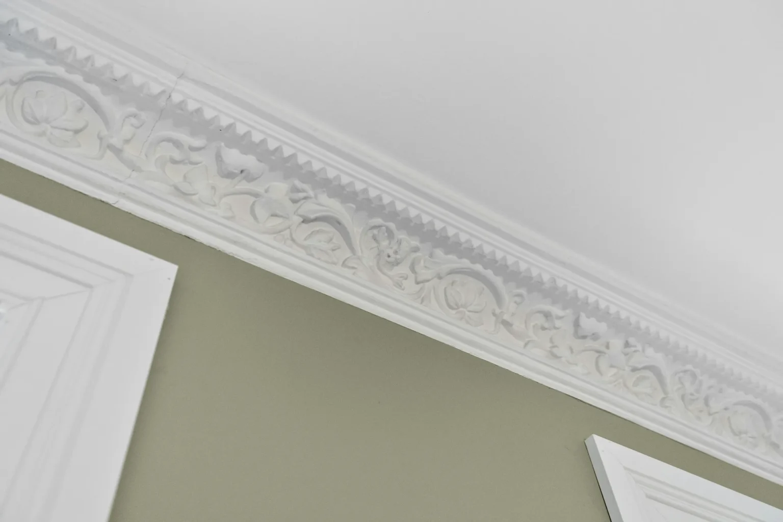 Corniche ornementée en plâtre blanc avec motif floral et vegetal, fixée au plafond au-dessus d'une porte blanche.