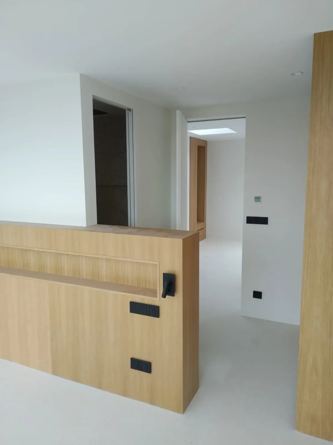 Meuble bas en bois clair avec interrupteurs noirs dans un couloir blanc menant à une autre pièce éclairée par un puits de lumière.