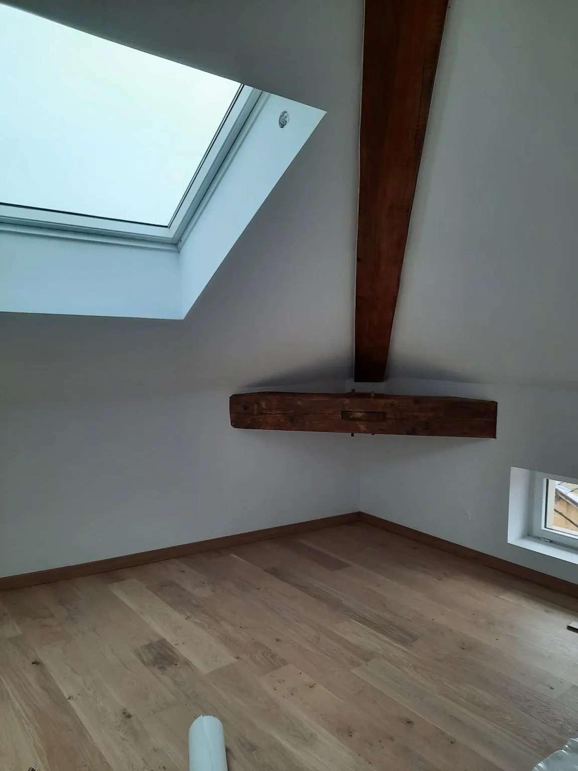 Poutres apparentes en bois sombre dans un grenier blanc avec parquet clair et un vélux.