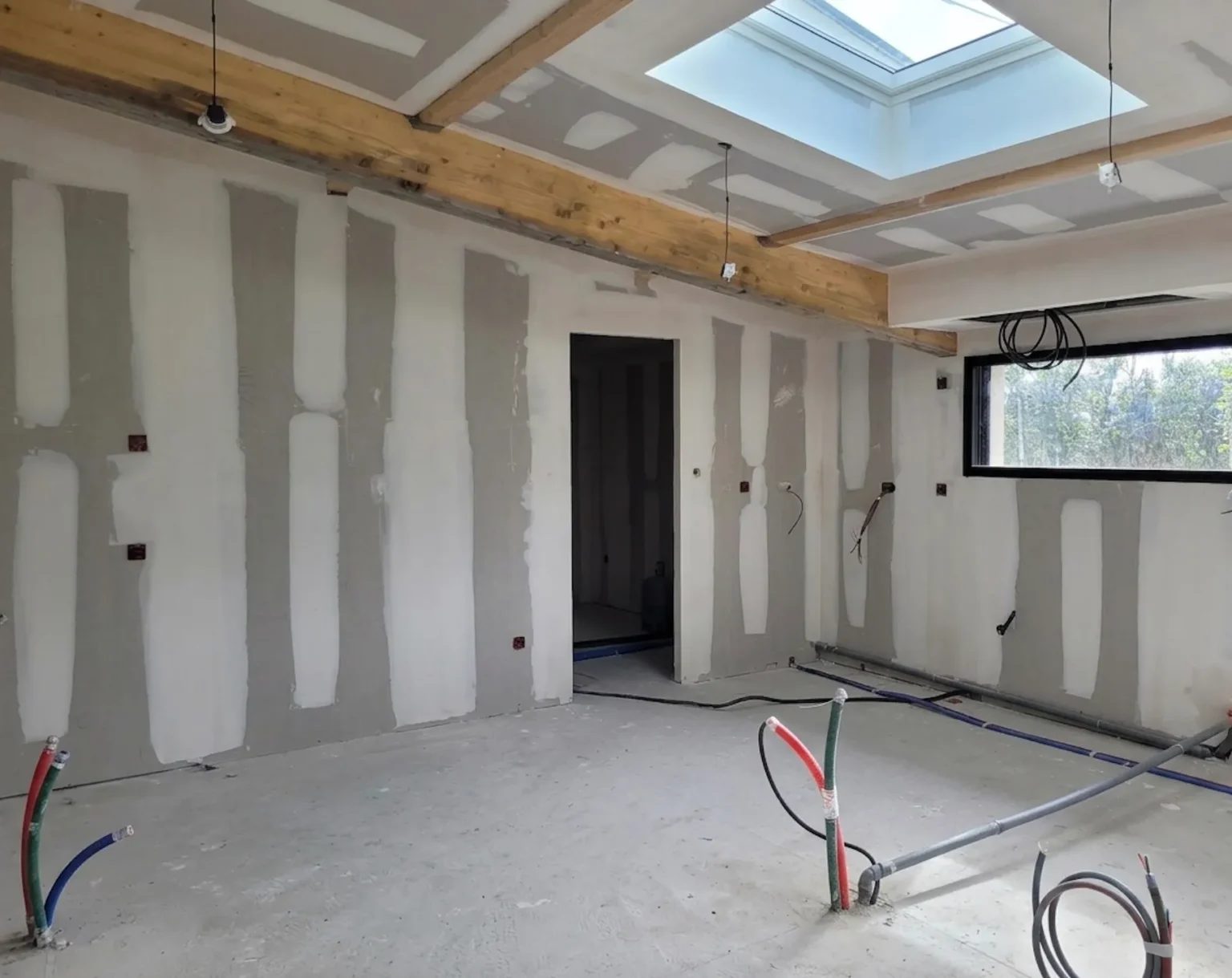 Intérieur d'une maison en construction, murs en plaques de plâtre, poutres apparentes et fenêtre de toit.