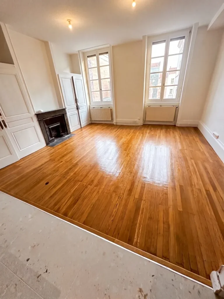 Chambre avec parquet en bois brillant, deux grandes fenêtres, un manteau de cheminée noir et des portes blanches.