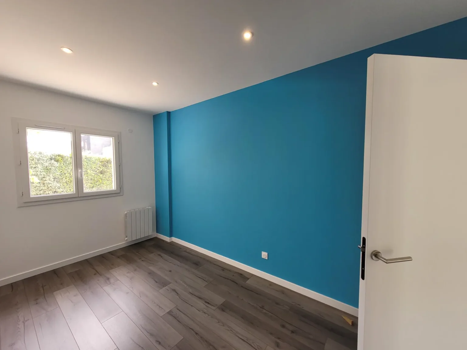 Chambre avec mur bleu vif, parquet gris, radiateur blanc et fenêtre donnant sur la verdure.