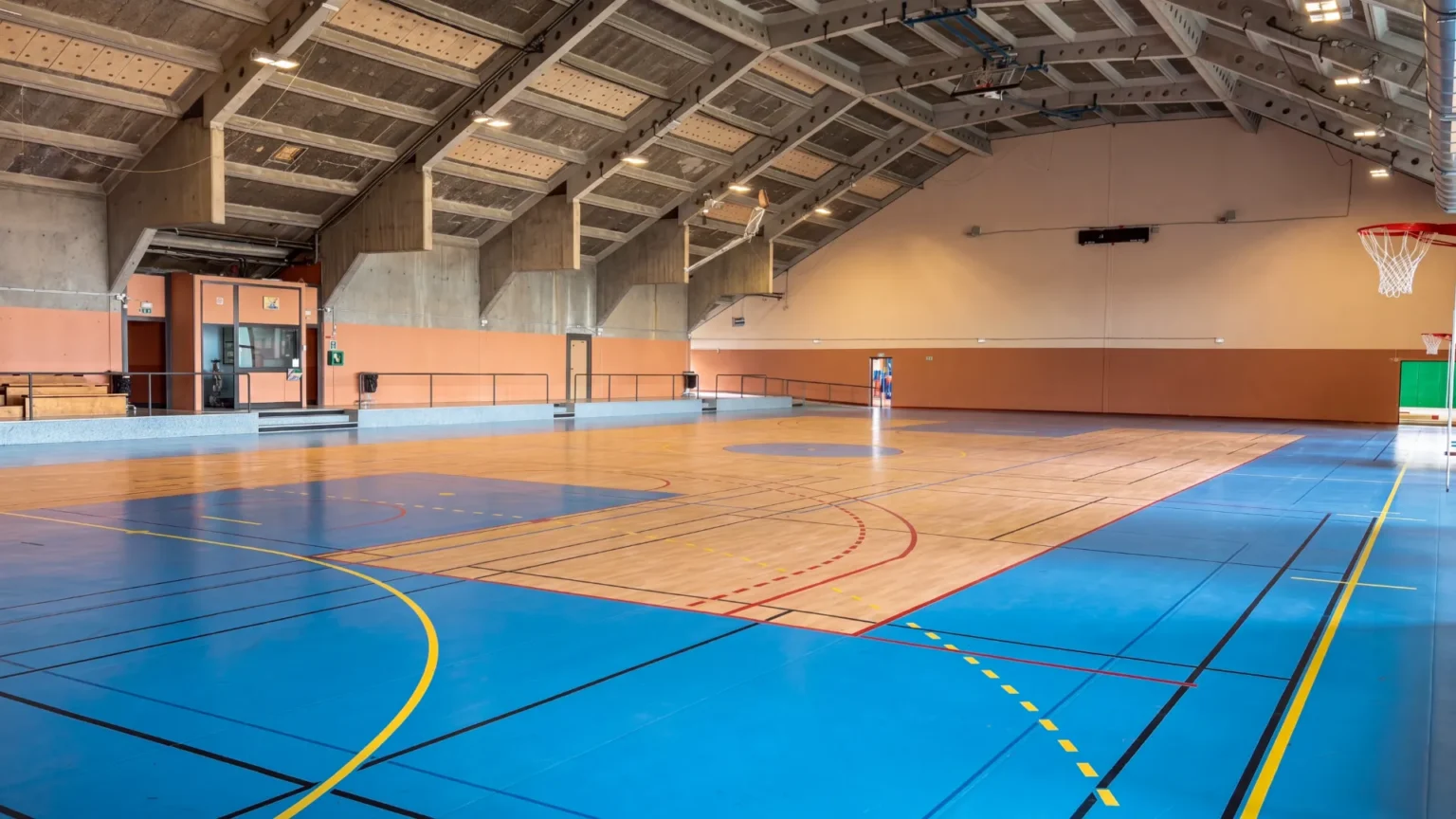 Intérieur d'une salle de sport avec un sol bleu et bois marqué pour plusieurs sports, un panier de basket est visible.