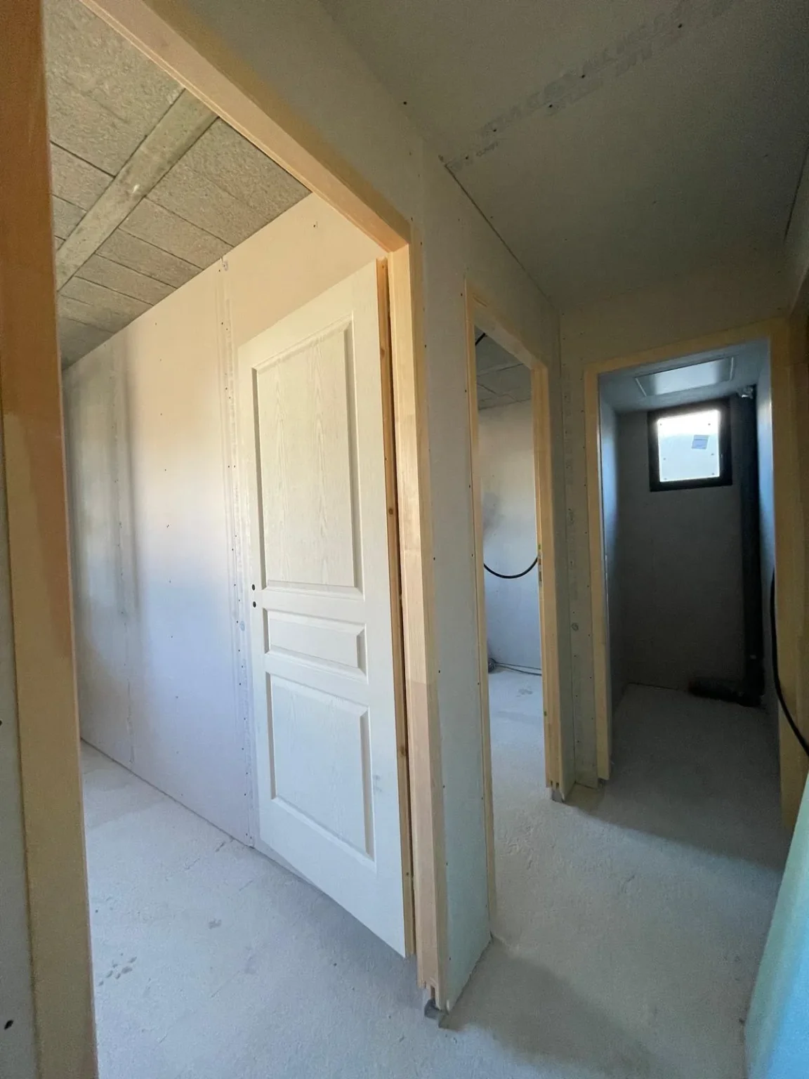 Couloir en travaux avec portes entrouvertes, cloisons en cours de finition et un petit fenêtre rectangulaire au fond.