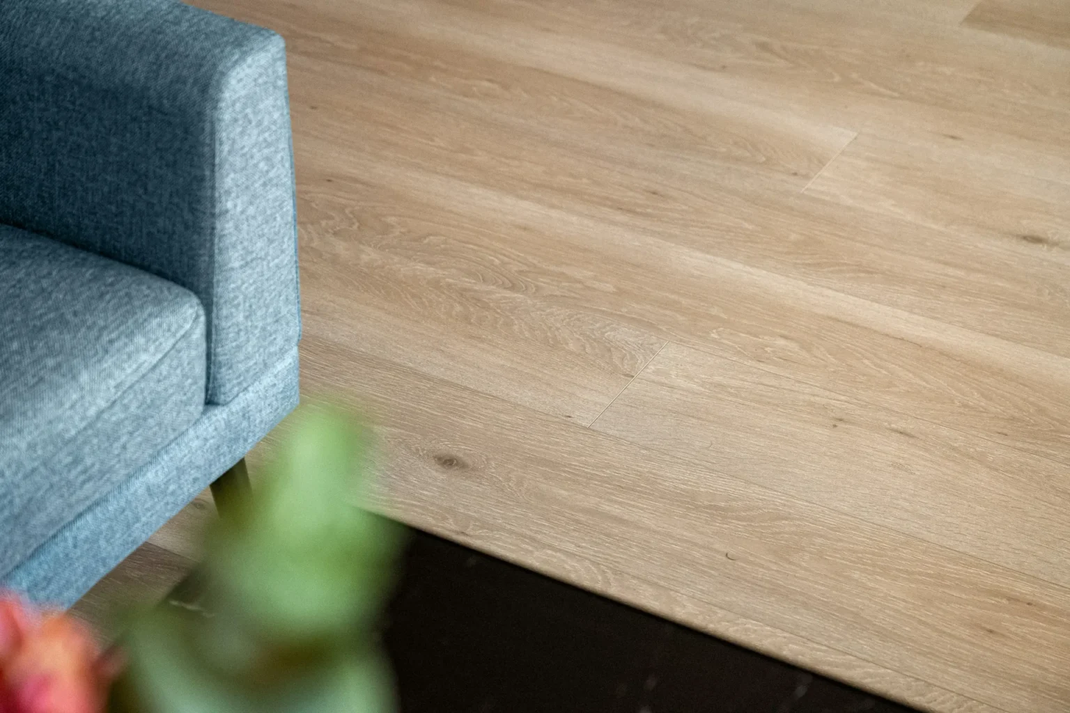 Gros plan sur un plancher aspect bois clair à côté d'un fauteuil bleu et d'une plante floue.