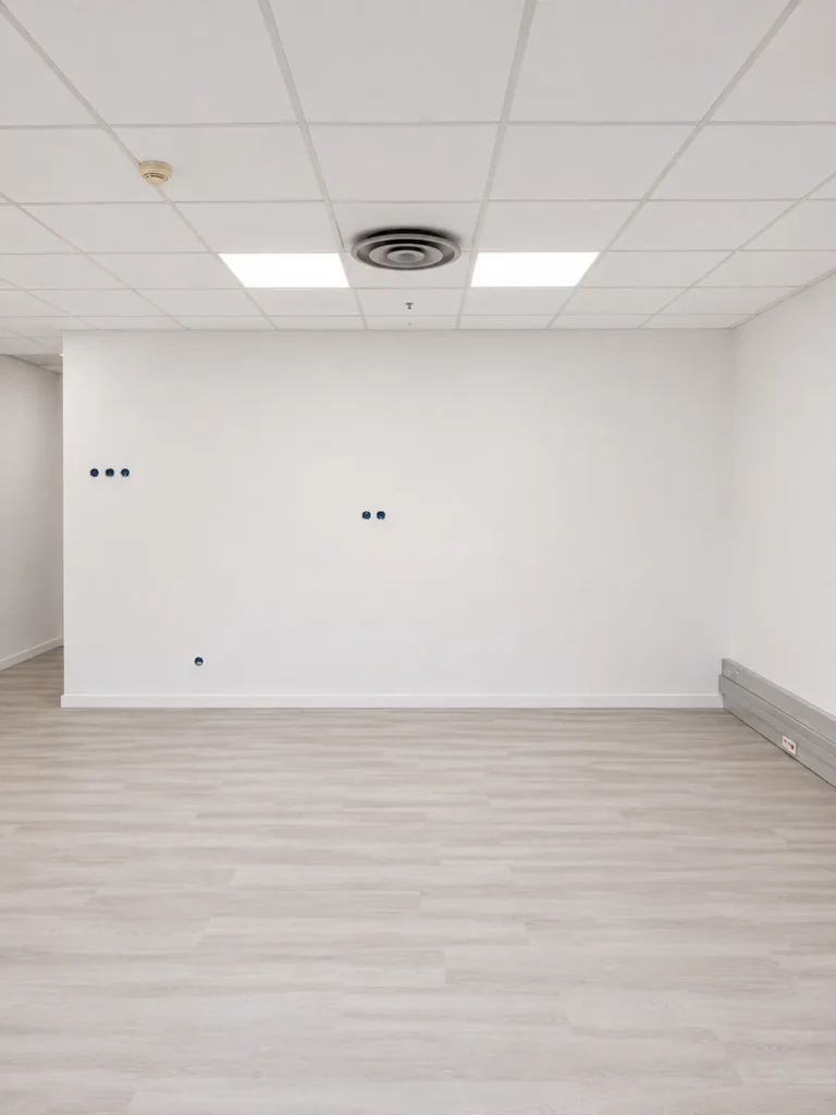 Intérieur d'une pièce vide avec sol stratifié imitation bois clair, murs blancs, faux plafond et éléments de ventilation.