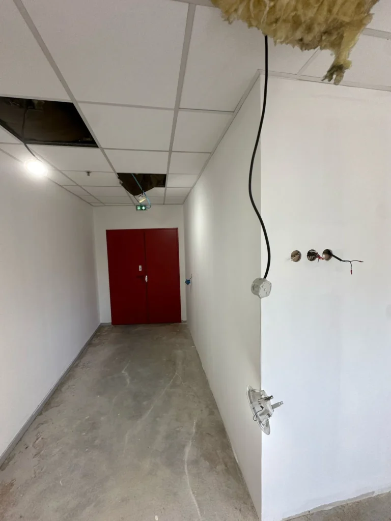 Couloir en construction avec portes rouges au fond, câbles apparents, prises électriques et isolation au plafond dégradé.