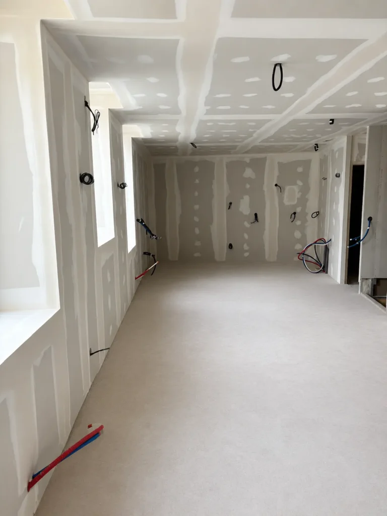 Chantier d'une pièce avec murs et plafond en plaques de plâtre non peints, des gaines électriques apparentes et des tuyaux installés.