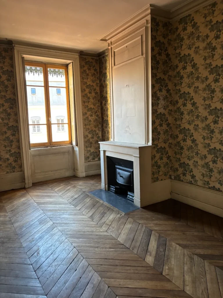 Intérieur de pièce avec parquet ancien en point de Hongrie, une cheminée blanche et un papier peint à motifs floraux.