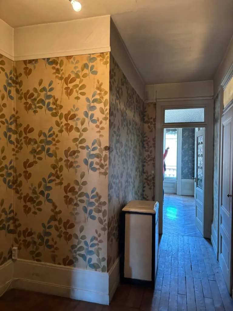 Couloir d'une maison ancienne avec un papier peint à motif de feuilles, un parquet en bois et des portes ouvertes sur d'autres pièces.