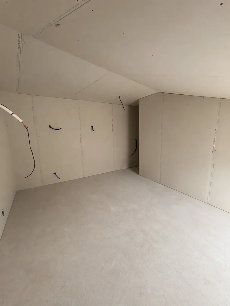 Intérieur d'une pièce en cours de construction, murs et plafond en plaques de plâtre, câblage électrique apparent.