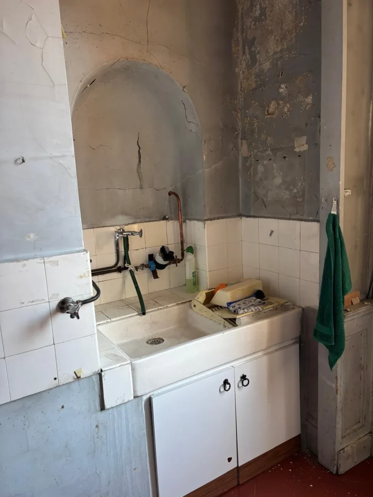 Ancien lavabo de cuisine dans une pièce nécessitant une rénovation, avec des murs fissurés et du carrelage défraîchi.